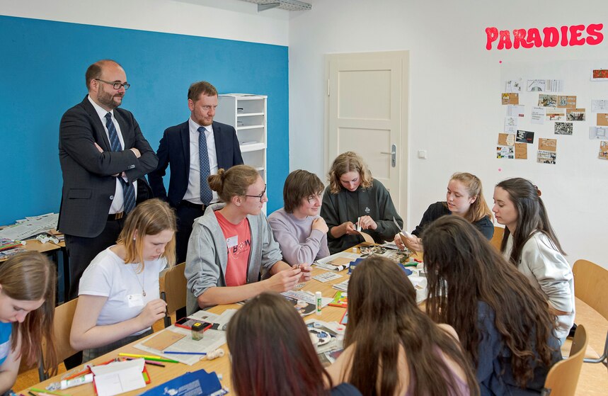 Personen sitzen sich gegenüber an einem Tisch und sind in die Arbeit vertieft. Zwei stehende Personen schauen zu.