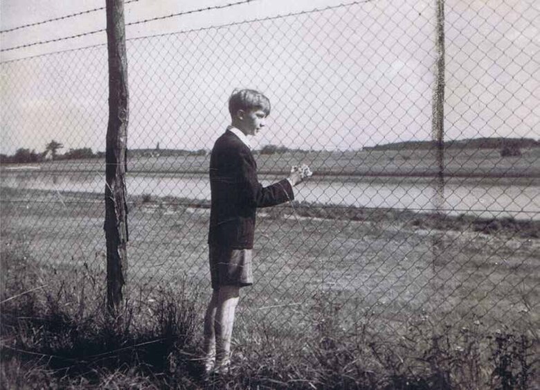 Eine Schwarz-Weiß-Aufnahme eines Jungen, der an einem Drahtzaun steht und in die Ferne blickt