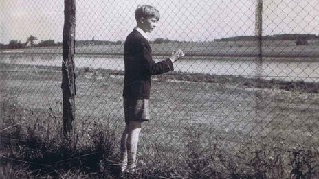 Eine Schwarz-Weiß-Aufnahme eines Jungen, der an einem Drahtzaun steht und in die Ferne blickt